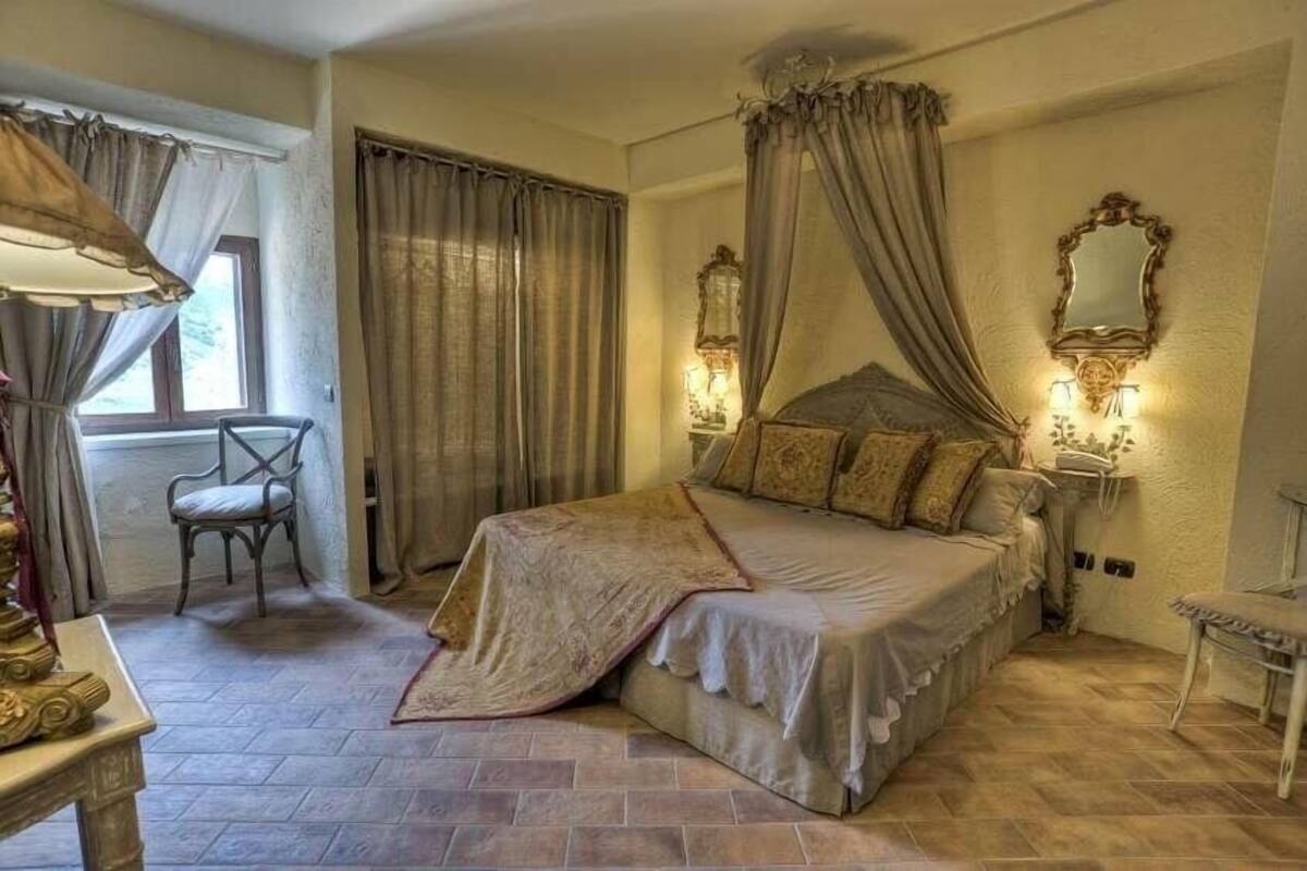 Slaapkamer met een hemelbed, een stoel, een nachtkastje met lamp en een raam met gordijnen.