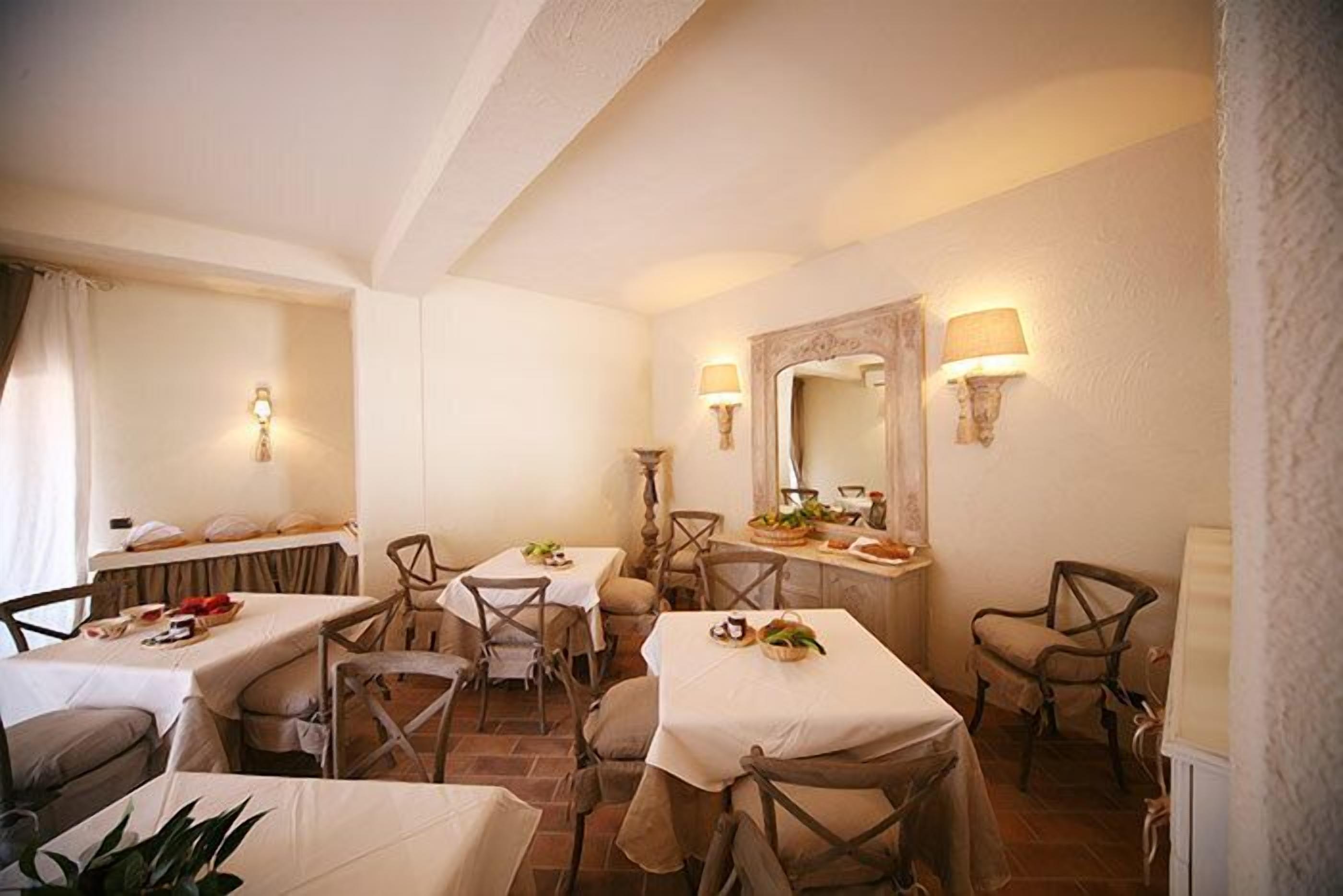Un comedor con cuatro mesas, cada una con mantel blanco y rodeada de sillas de madera. Hay lámparas fijadas a la pared a ambos lados de la habitación, y un espejo sobre la encimera.