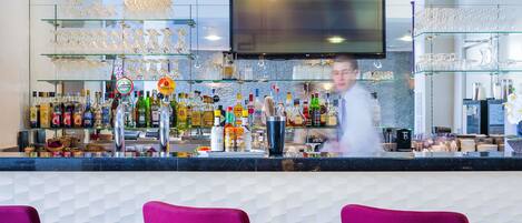 A bar area with a counter, shelves of bottles, glasses, and a TV screen.