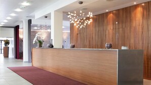 Un comptoir d’accueil avec un panneau en bois en arrière-plan, un lustre moderne et un tapis rouge menant à l’entrée.