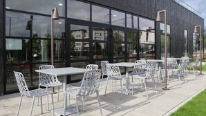 Un espace extérieur aménagé avec des tables et des chaises en métal blanc, une façade de bâtiment moderne et une vue dégagée sur les environs.