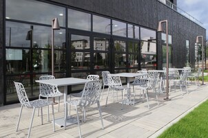 Un espacio al aire libre con mesas y sillas de metal blanco, fachada de un edificio moderno y vistas despejadas del entorno.