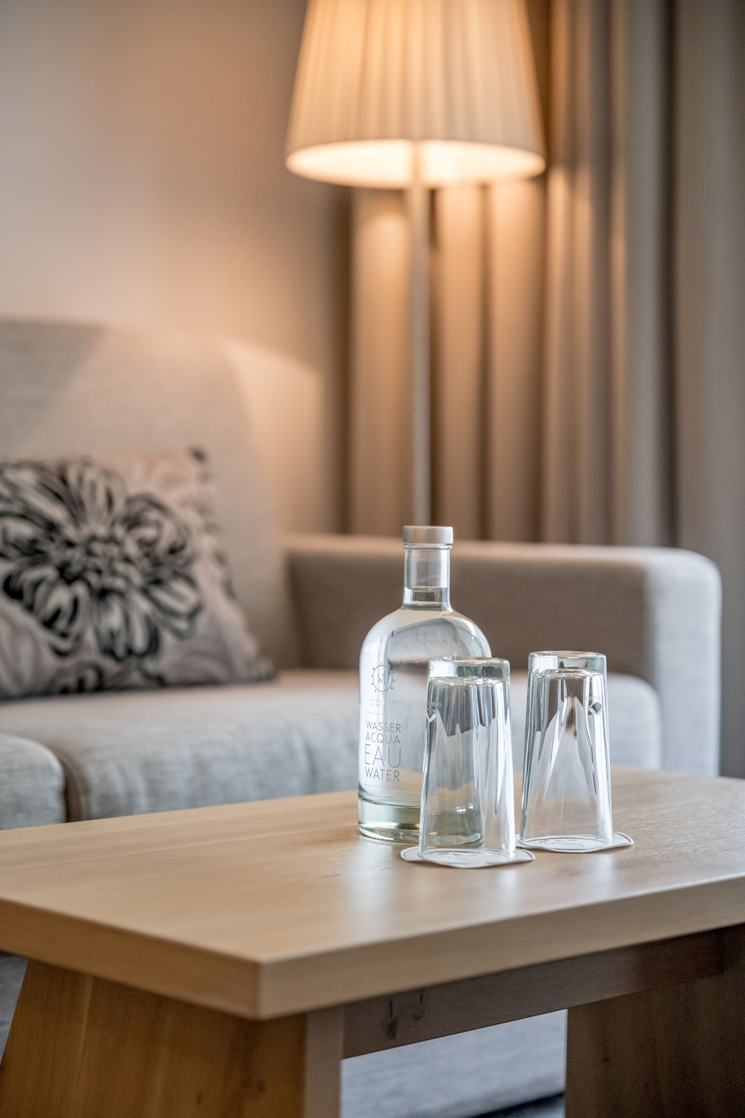 A wooden table with a clear bottle and two glasses on it, set on a sofa with a decorative pillow and a lamp in the background.
