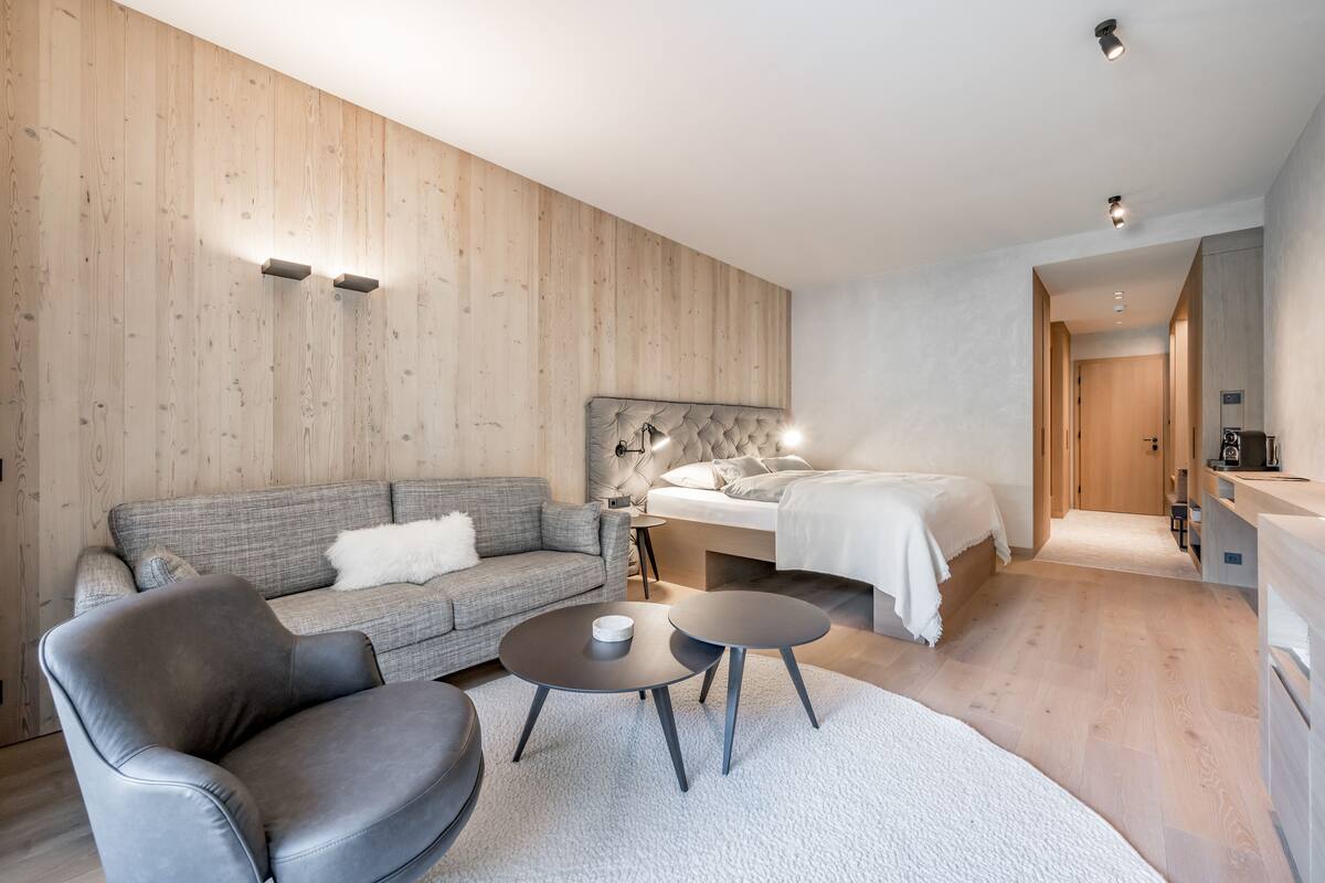 Une chambre d’hôtel moderne avec un mur en bois, un canapé gris, un lit, une table de chevet et un petit bureau avec une lampe.