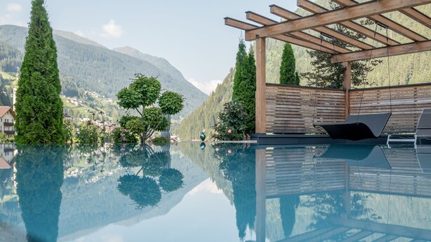 Vista despejada de una piscina con una pérgola de madera y sillones de descanso, rodeada de exuberante vegetación y montañas al fondo.