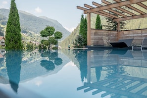 A clear view of a swimming pool with a wooden pergola and lounge chairs, surrounded by lush greenery and mountains in the background.
