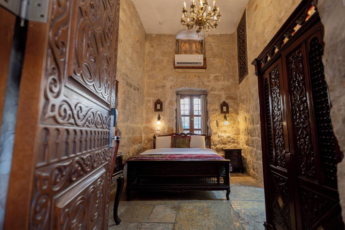 A bedroom with a stone wall, a wooden bed, a chandelier, and a window with curtains.