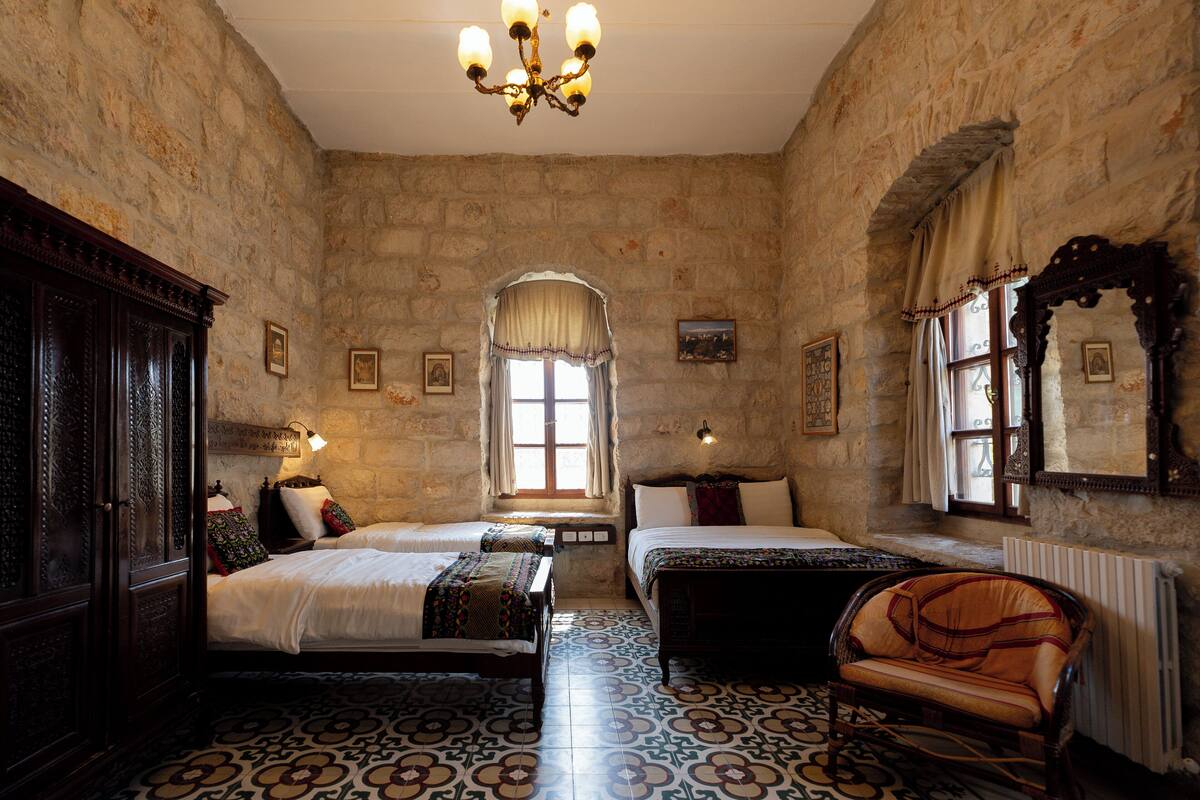 A room with two beds, a chair, a chandelier, and stone walls.