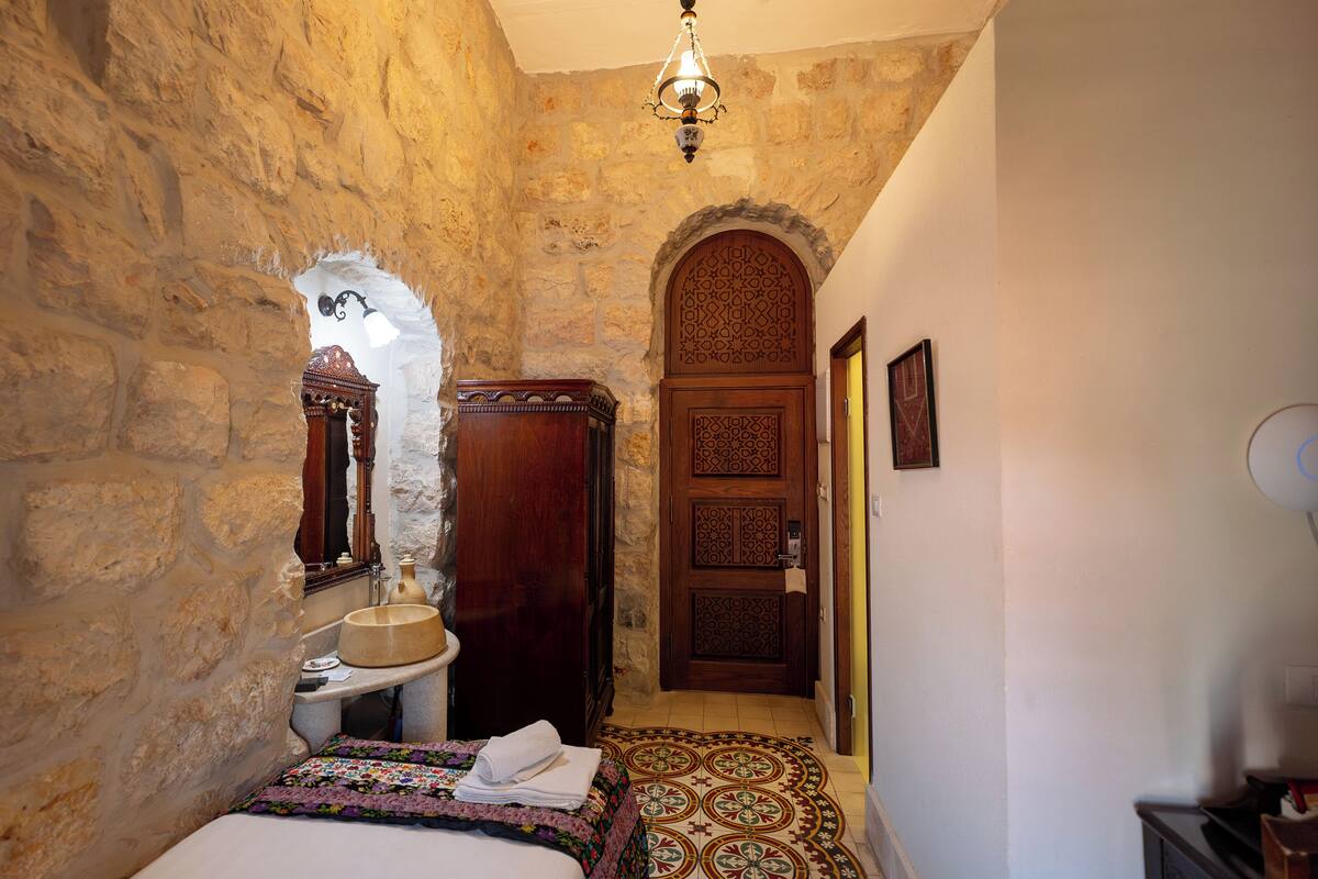 A narrow hallway with stone walls, a wooden door with intricate patterns, a bed with a colorful blanket, and a small sink with a mirror.