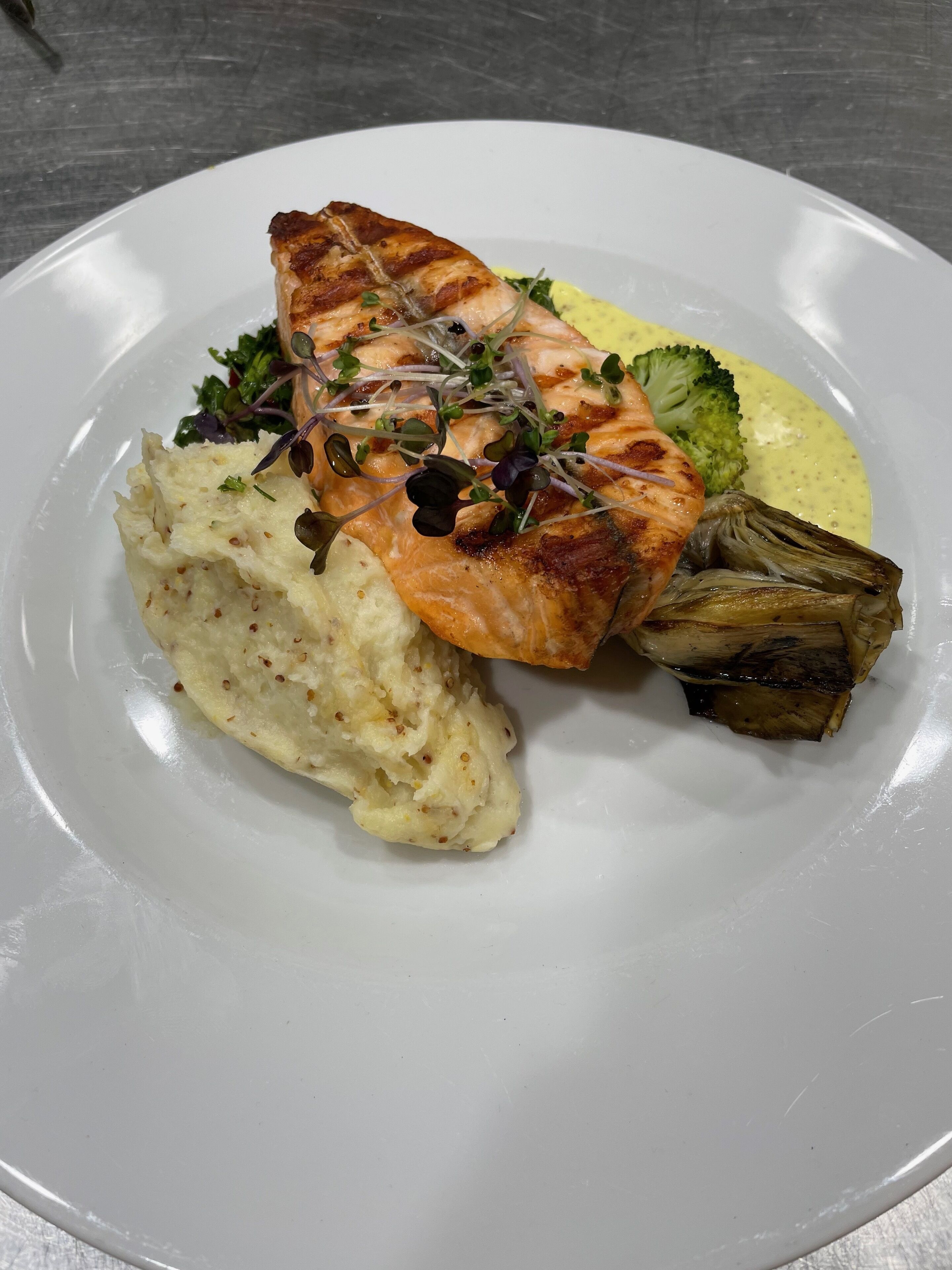 Een bord met gegrilde zalm, gepureerde aardappelen, broccoli en gegrilde artisjok.