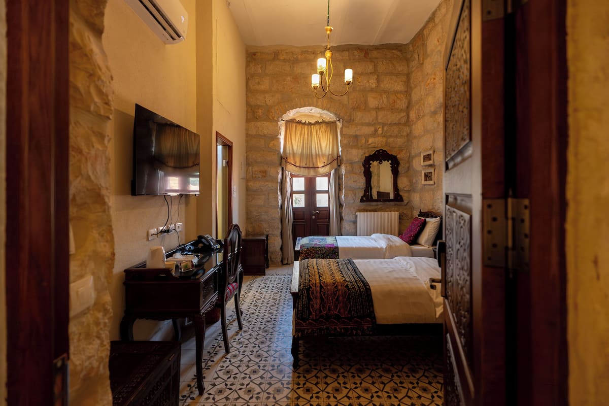 A traditional room with a stone wall, a flat-screen TV mounted on the wall, a chandelier, a bed with a patterned bedspread, and a wooden dresser.