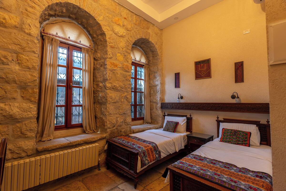 A room with stone walls, a bed, a bench, and two windows with curtains.