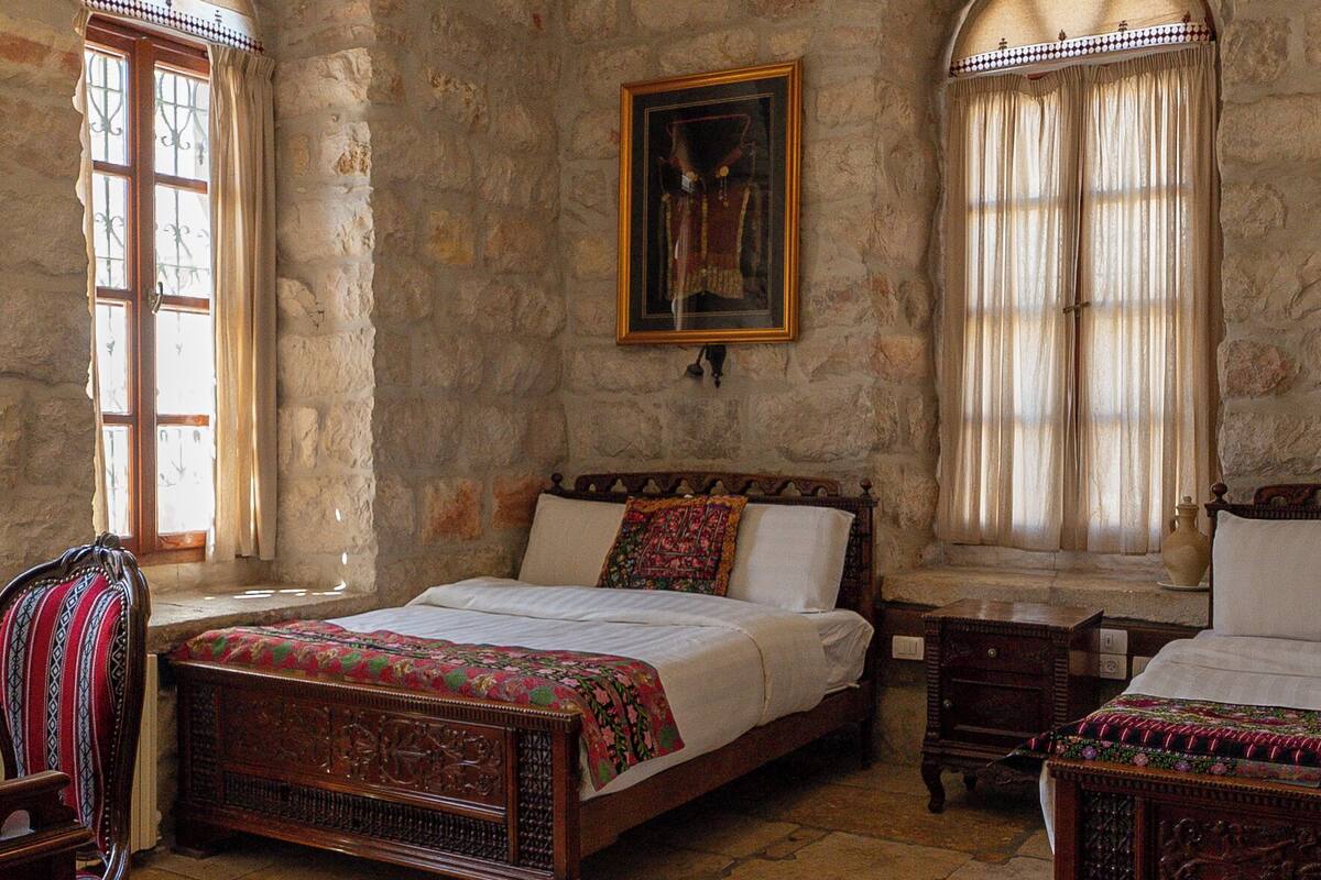 A traditional room with stone walls, a bed with a wooden headboard, two armchairs, a table, and a hanging light fixture.