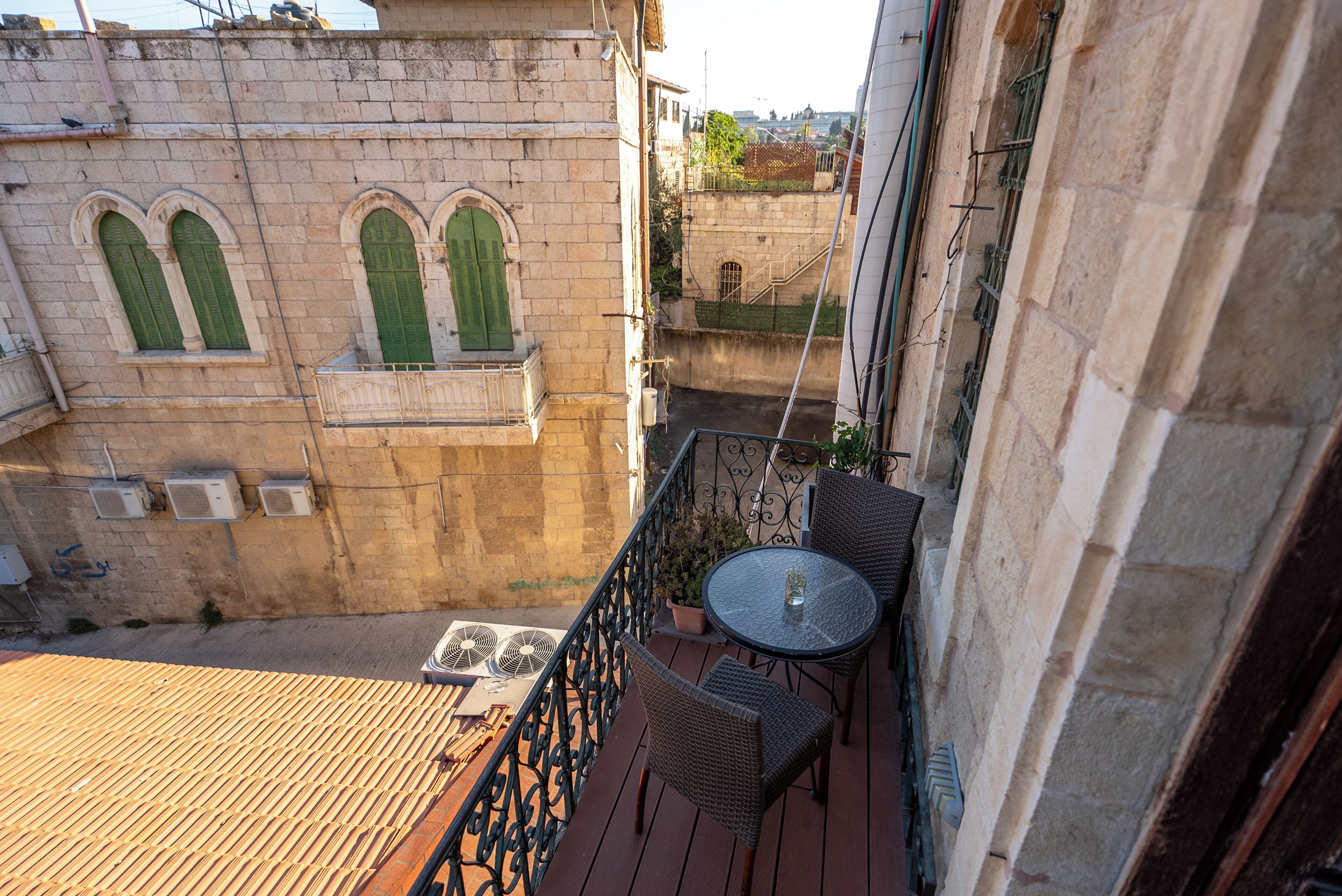 A balcony with wicker chairs and a small table, overlooking a courtyard with a stone wall and arched windows.
