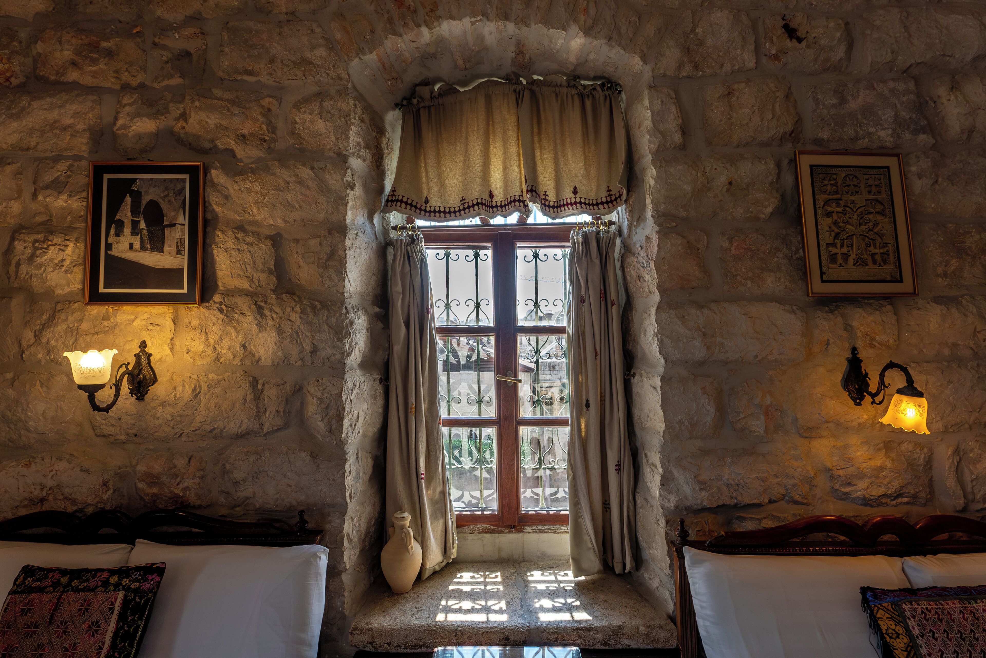 A room with a stone wall, a window with curtains, a bed with a headboard, and wall-mounted light fixtures.