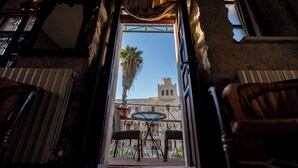 Interieur van een kamer met balkon, een tafel en stoelen, uitzicht op een palmboom en een gebouw op de achtergrond.