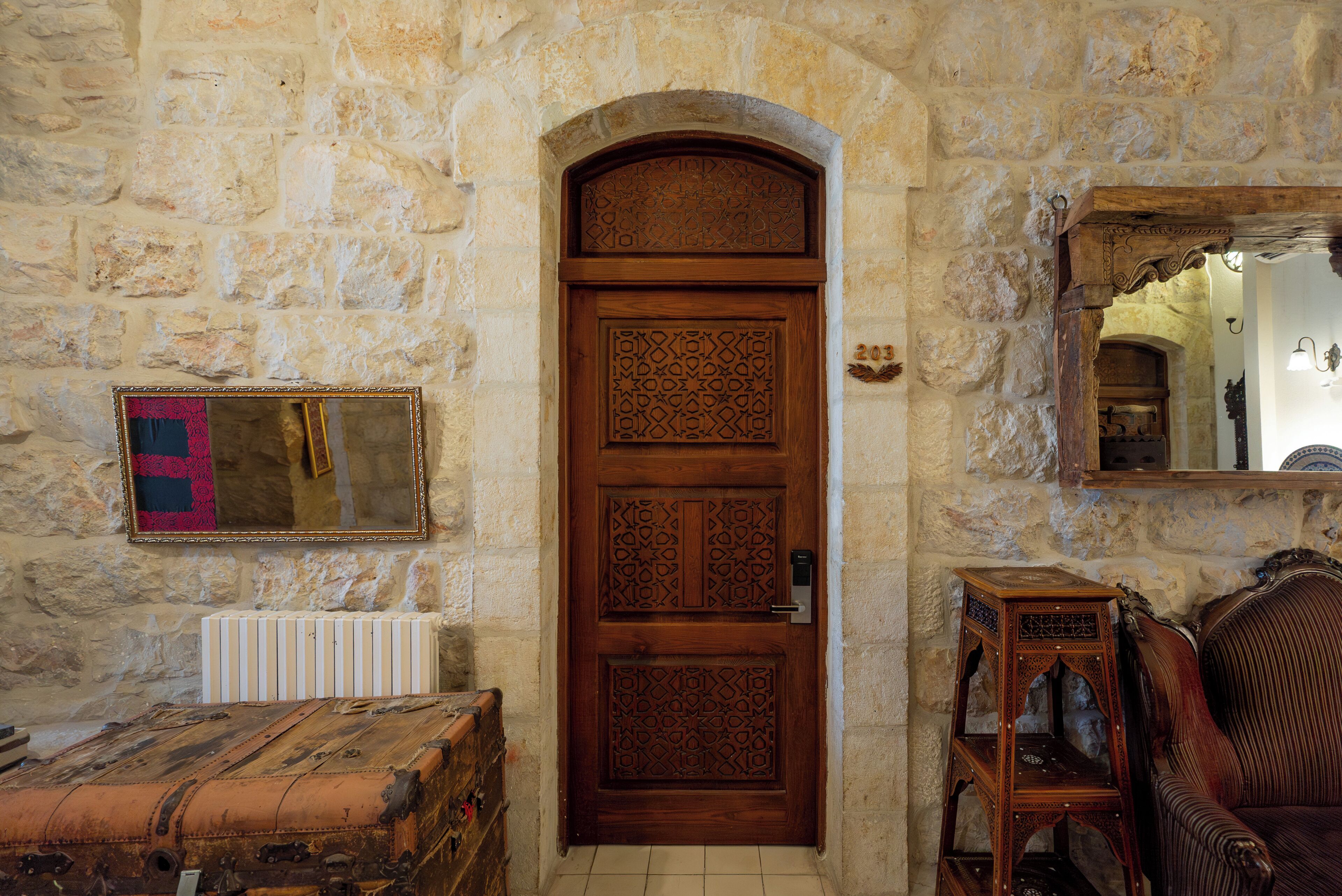 A stone-walled room with a wooden door, a radiator, a mirror, and a wooden chest.