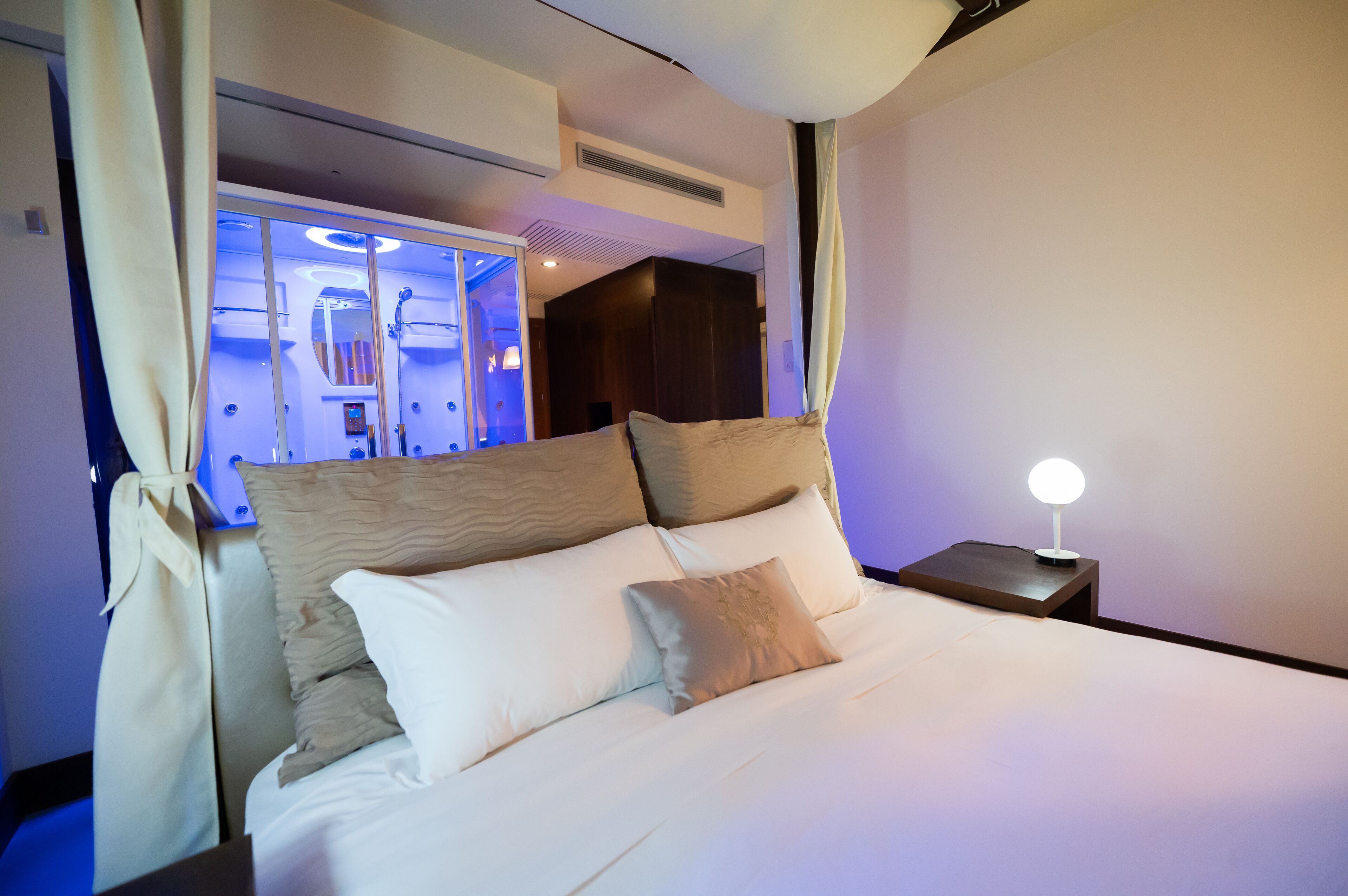 Habitación de hotel con una cama grande, una mesita de noche con lámpara y vistas a un baño con ducha y espejo.