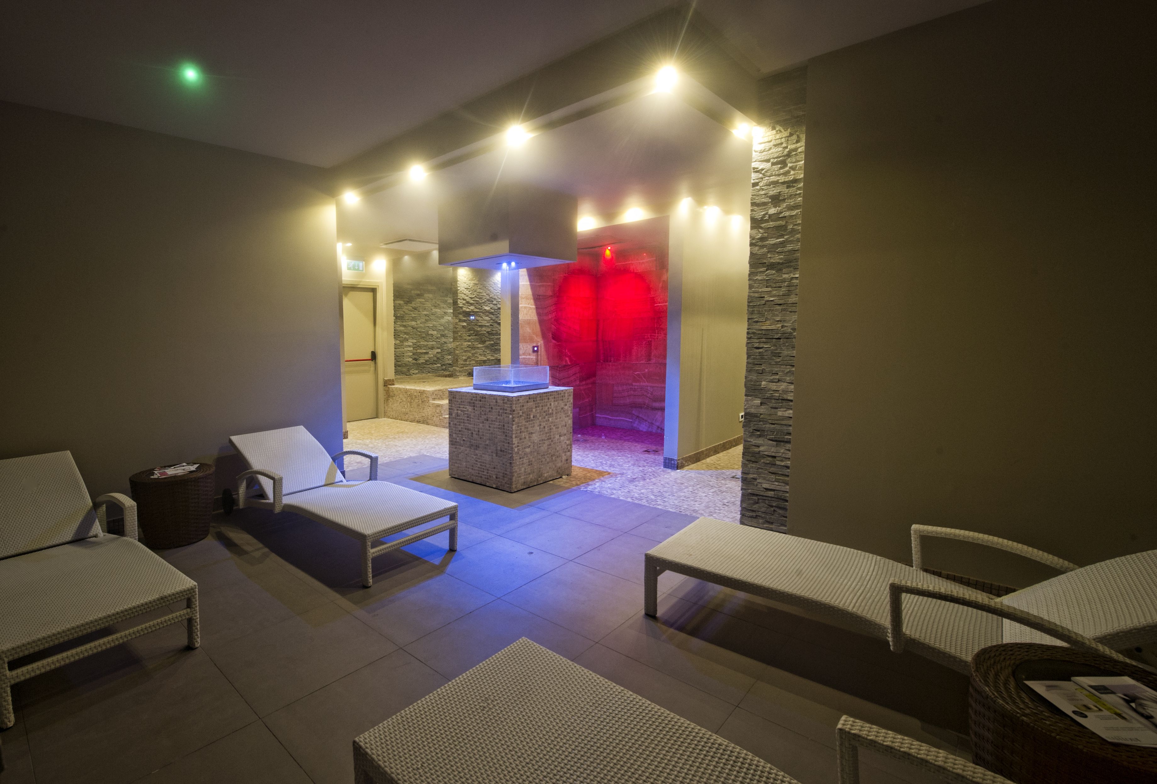 A spa area with white lounge chairs, a massage table, and a stone wall with a red light.