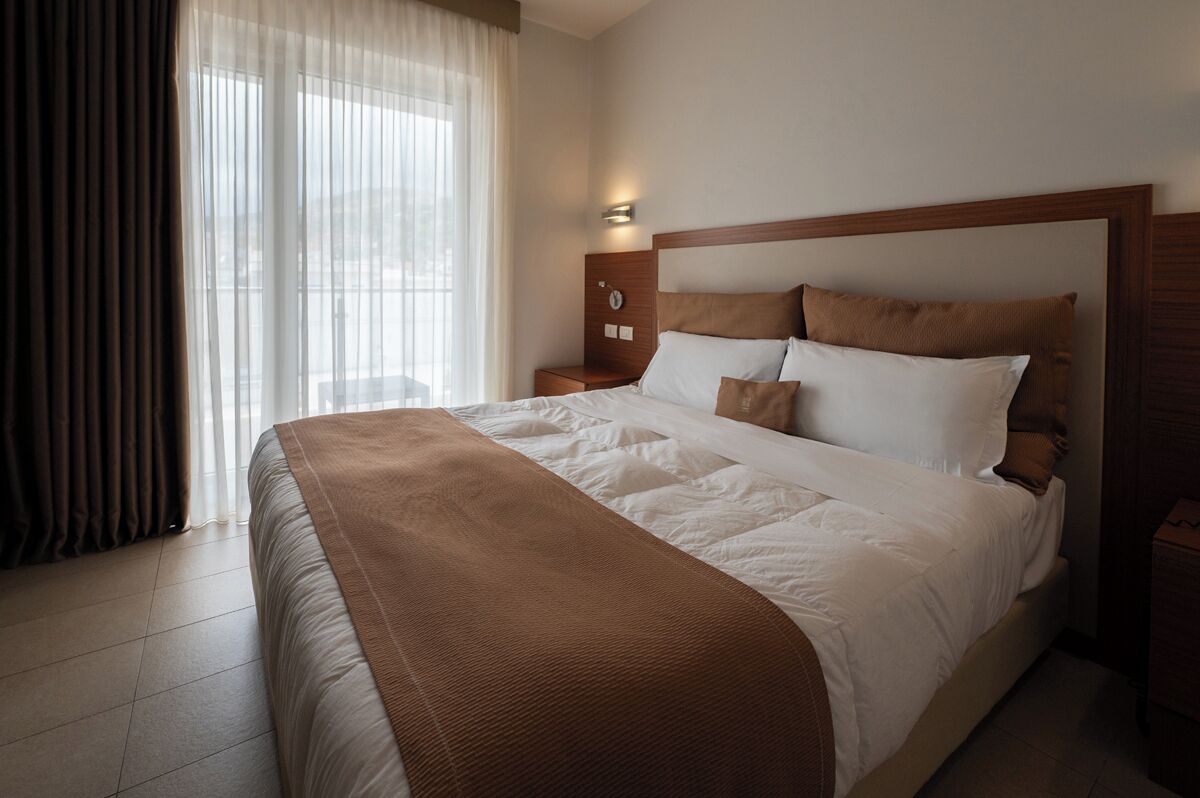 A hotel room with a large bed, a bedside table with a clock, and a window with sheer curtains.
