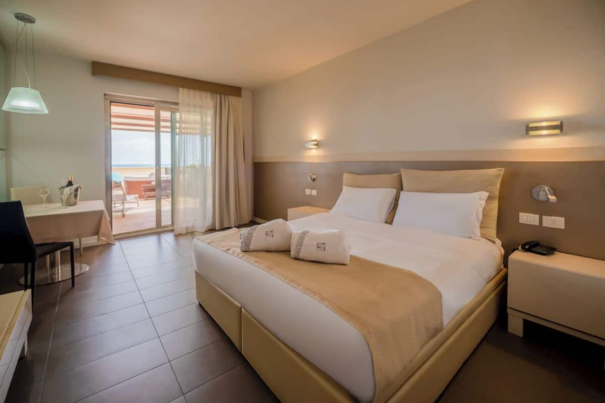 A hotel room with a bed, bedside tables, a desk with a chair, and a view of the beach through a sliding door.