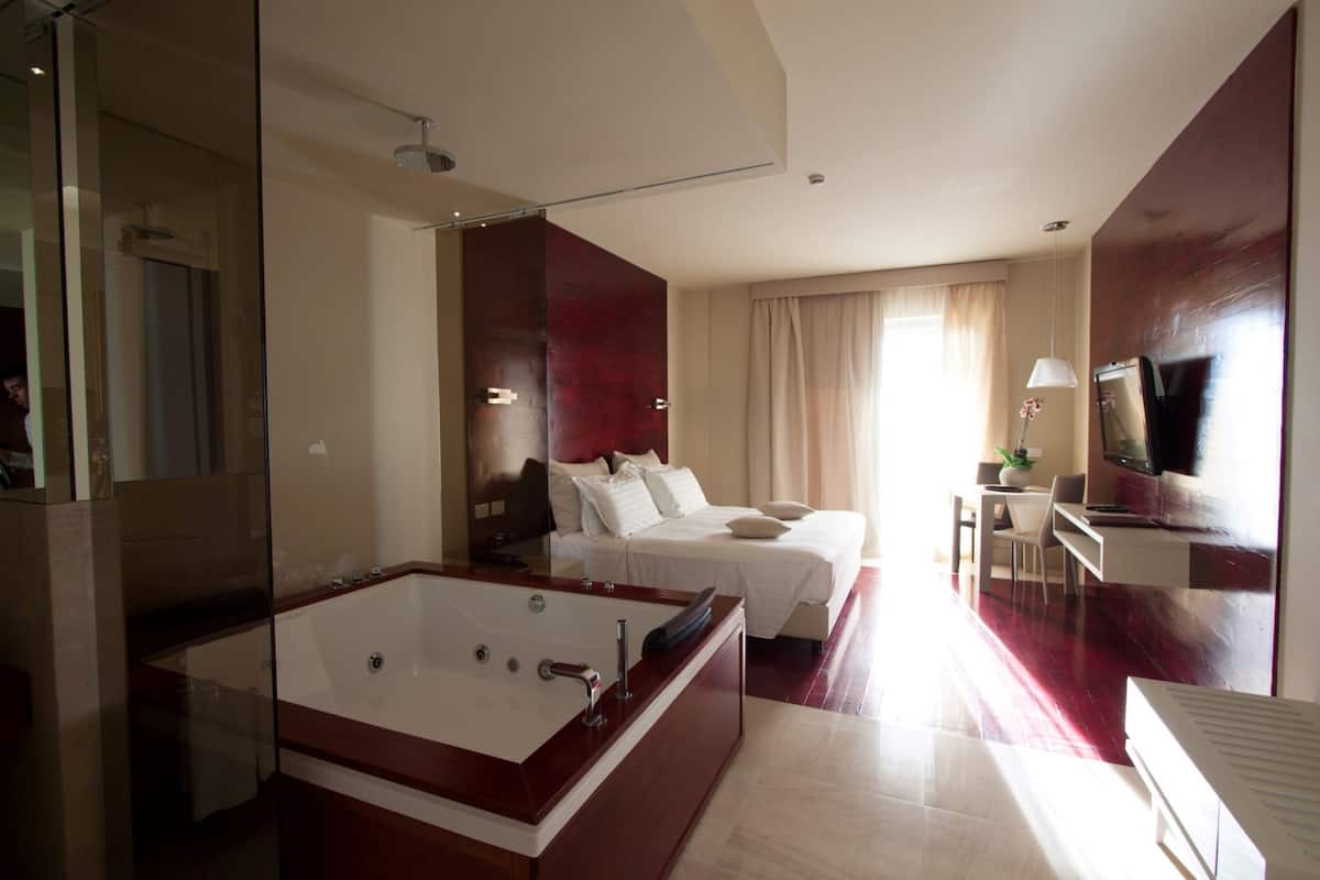 A modern hotel room with a large bathtub, a bed with white linens, a desk with a chair, and a television.