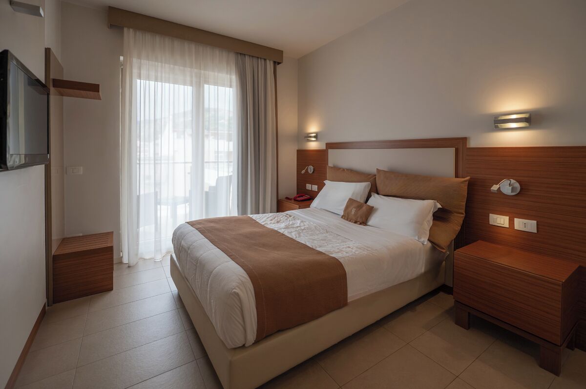 Ein Hotelzimmer mit einem großen Bett, Nachttischen, einem Fernseher und einem Fenster mit Vorhängen.