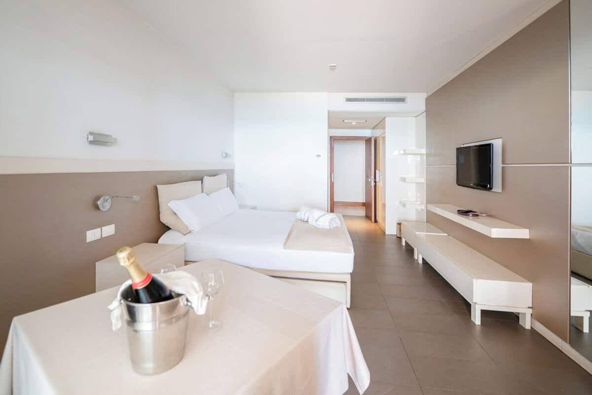 A modern hotel room with a bed, a TV, a minibar, and a table with a bottle of champagne.