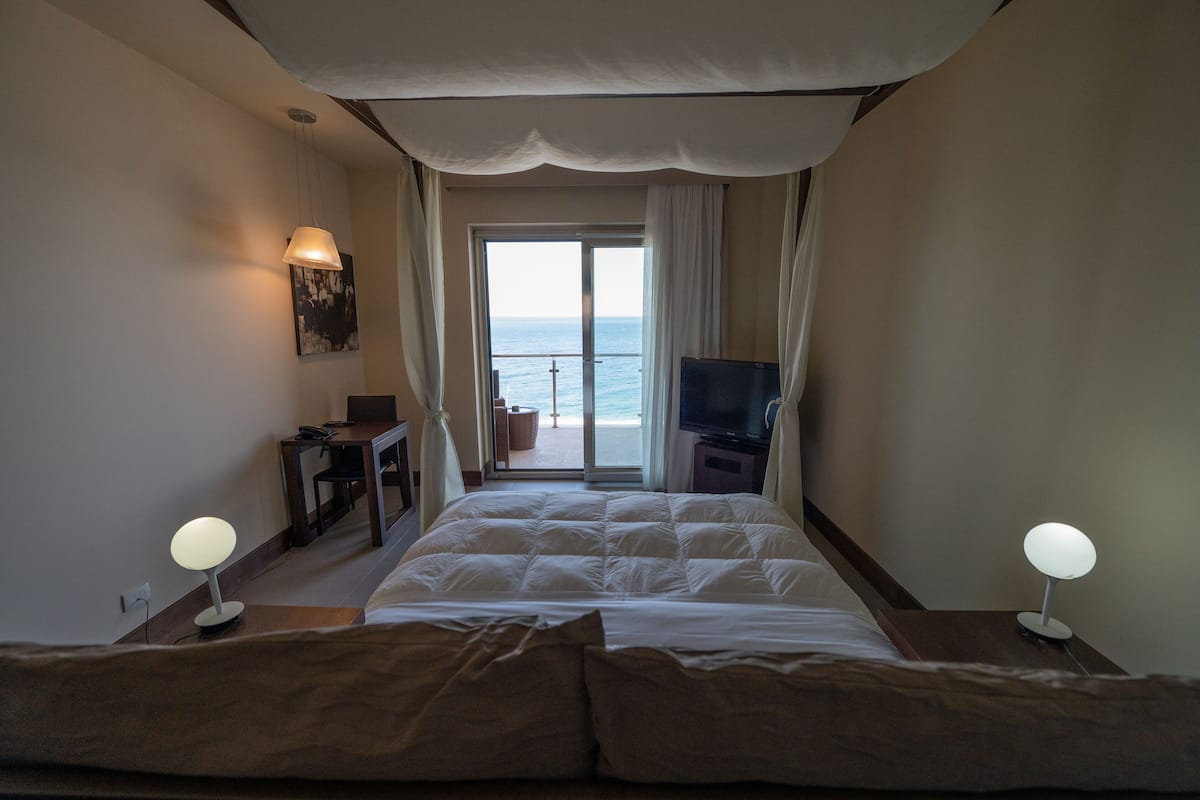 A hotel room with a large bed, a desk, a television, and a view of the sea through a sliding door.