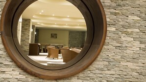 Un miroir rond reflète un espace d’accueil avec des meubles en osier et un mur en pierre.