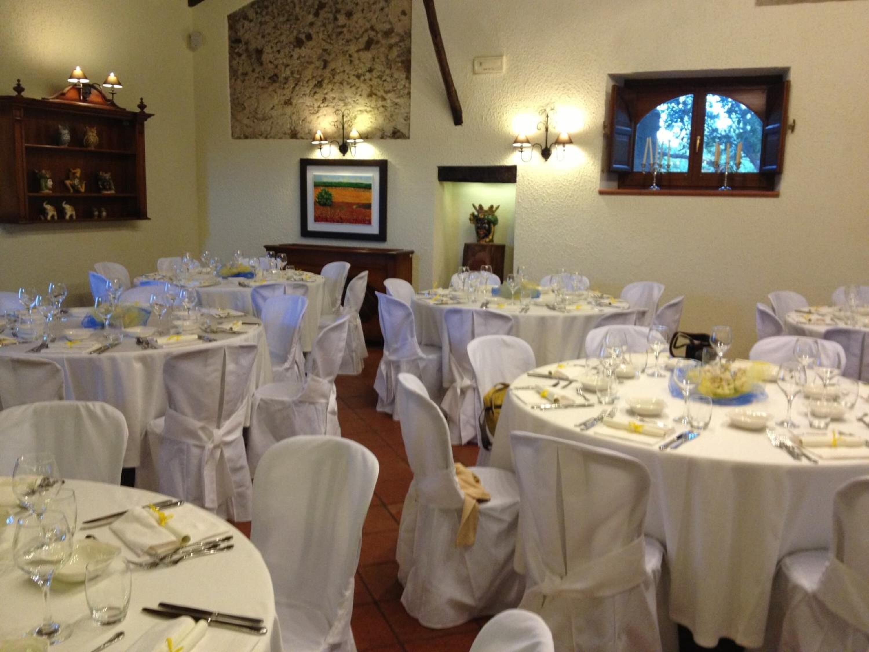 Une salle de banquet avec des tables rondes dressées pour le repas, des chaises blanches et un mur en pierre orné d’un tableau encadré.