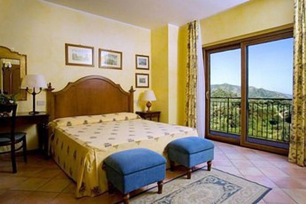 A bedroom with a bed, two blue ottomans, a nightstand, a lamp, and a large window with a view of mountains.