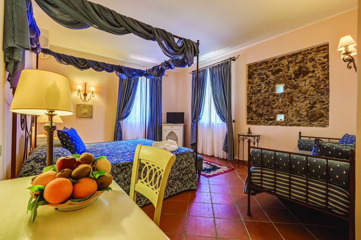 A hotel room with a bed, a sofa, a dining table with fruit, curtains, and a stone wall.
