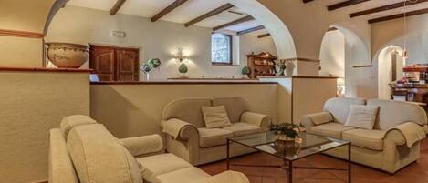 Una sala de estar acogedora con una mesa de centro de vidrio, sofás beige y una cocina visible a través de un arco.