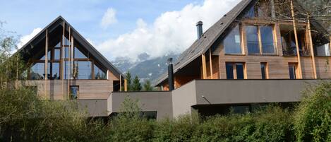 A modern two-story house with large windows and a balcony, surrounded by greenery and mountains in the background.