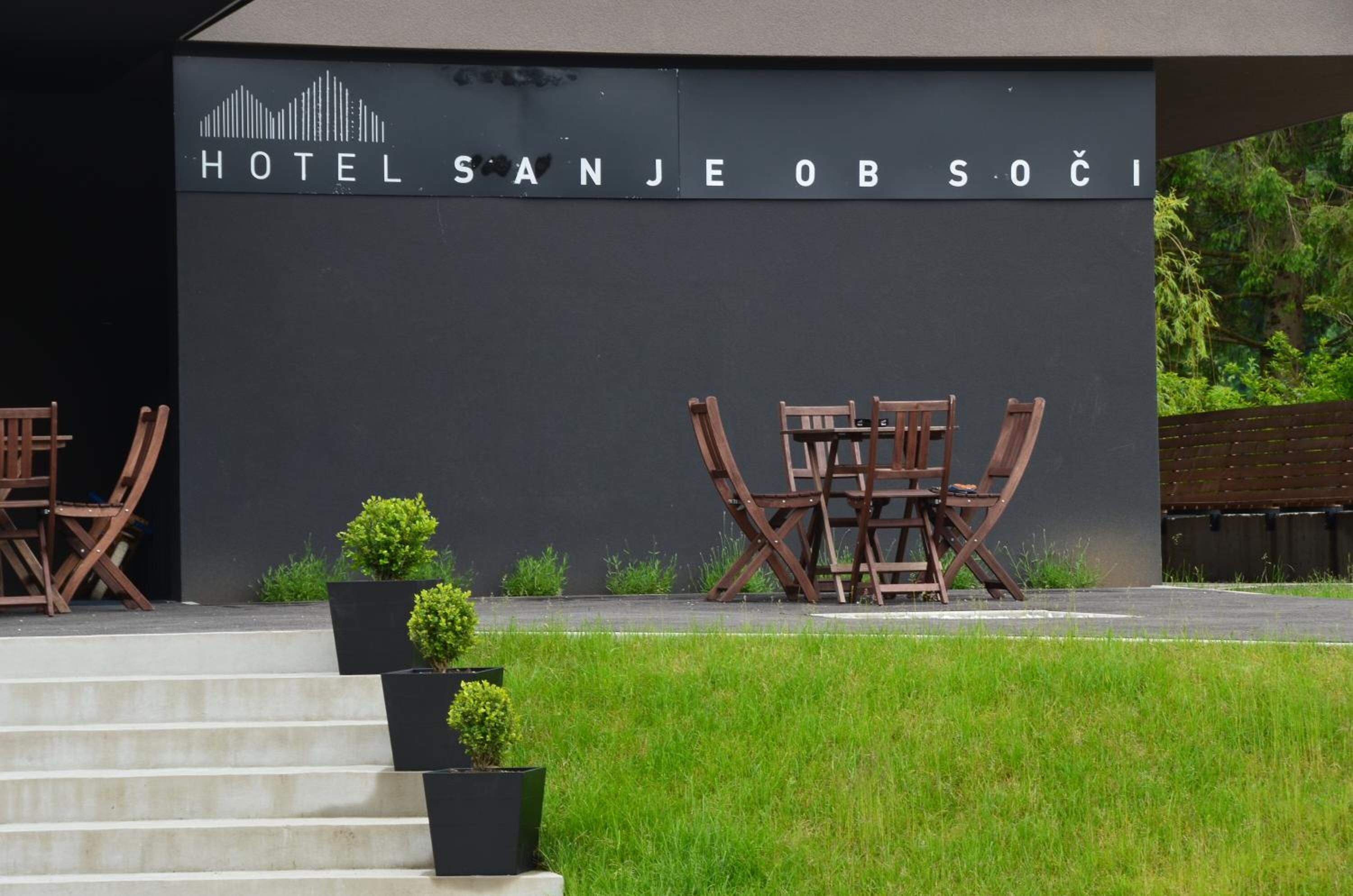 Entrada de un hotel con un letrero que dice "HOTEL SANJE OB SOĆI" y área de estar al aire libre con sillas y mesas de madera.