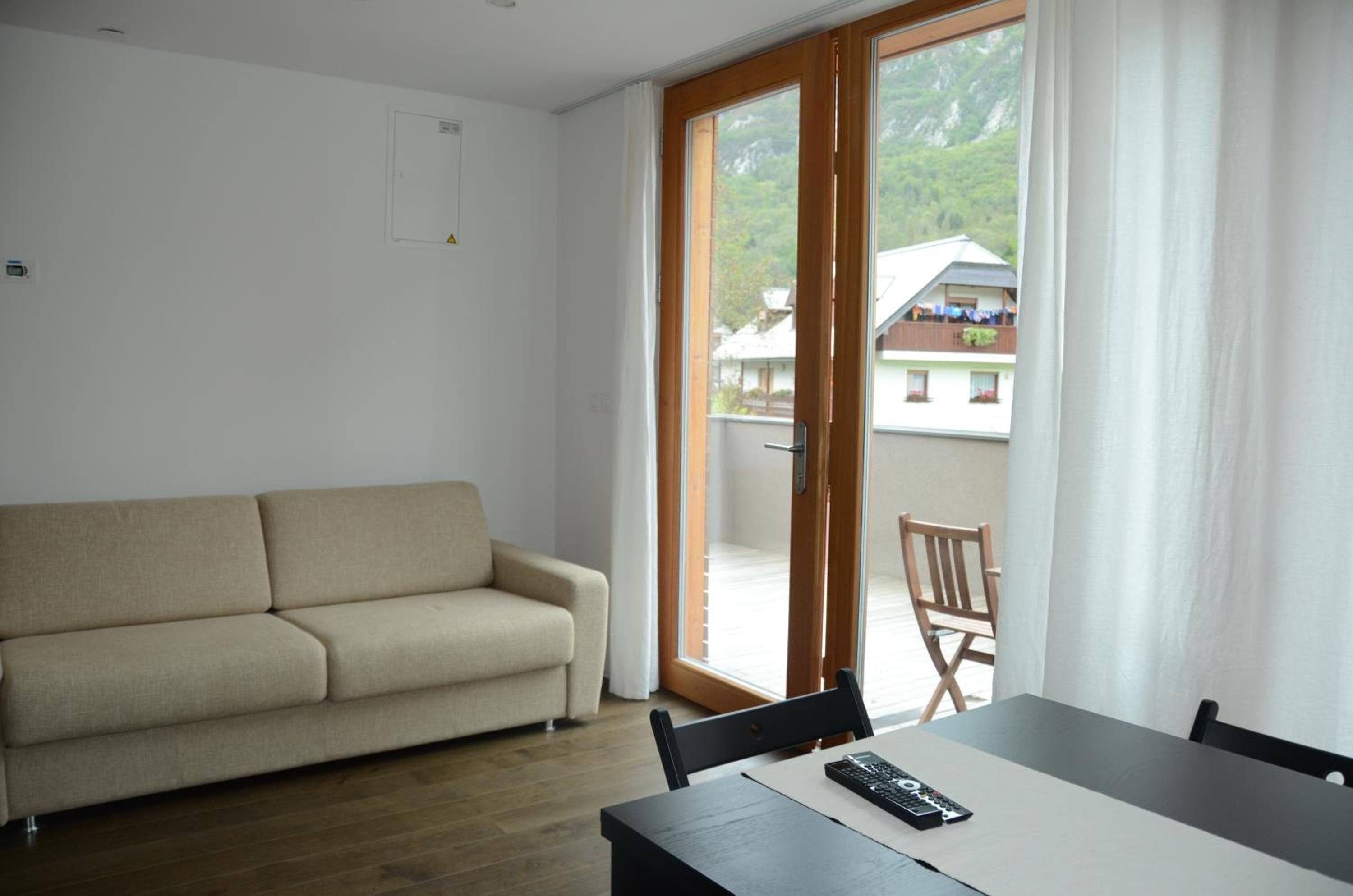 Een kamer met een beige bank, een eettafel met stoelen, een afstandsbediening en een schuifdeur die uitkomt op een buitenruimte met uitzicht op huizen en groen.