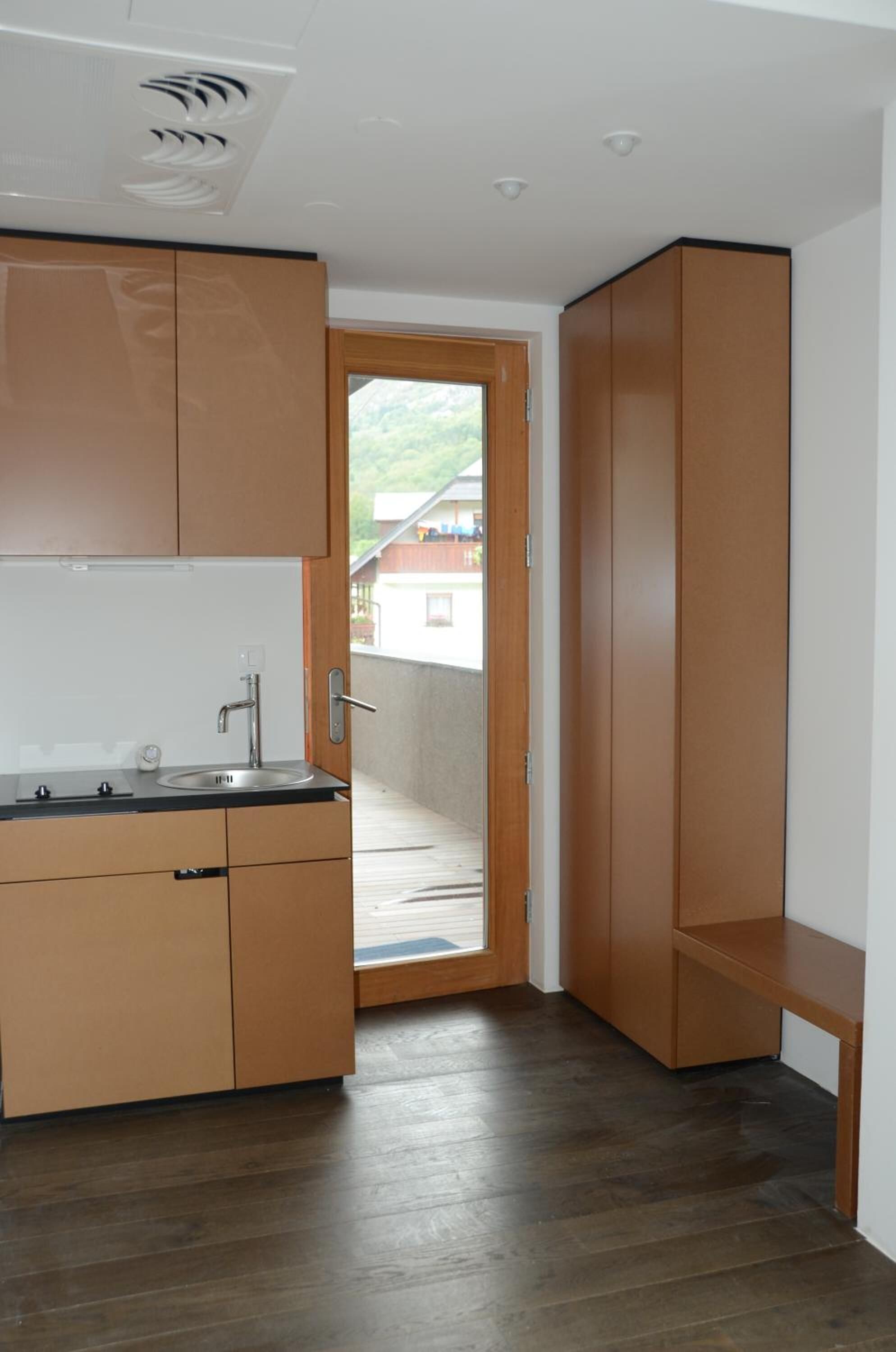 Cocina compacta con armarios de madera, un lavamanos y una puerta que da a un balcón.