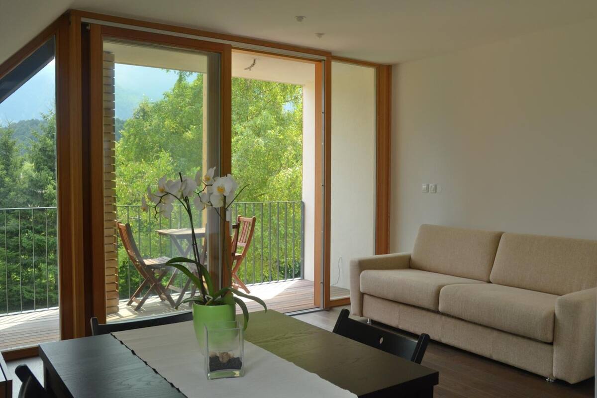 Un salon moderne avec un canapé, une table à manger agrémentée d’un vase de fleurs et un balcon offrant une vue sur la verdure.