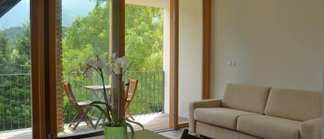 A modern living room with a sofa, a dining table with a vase of flowers, and a balcony with a view of greenery.