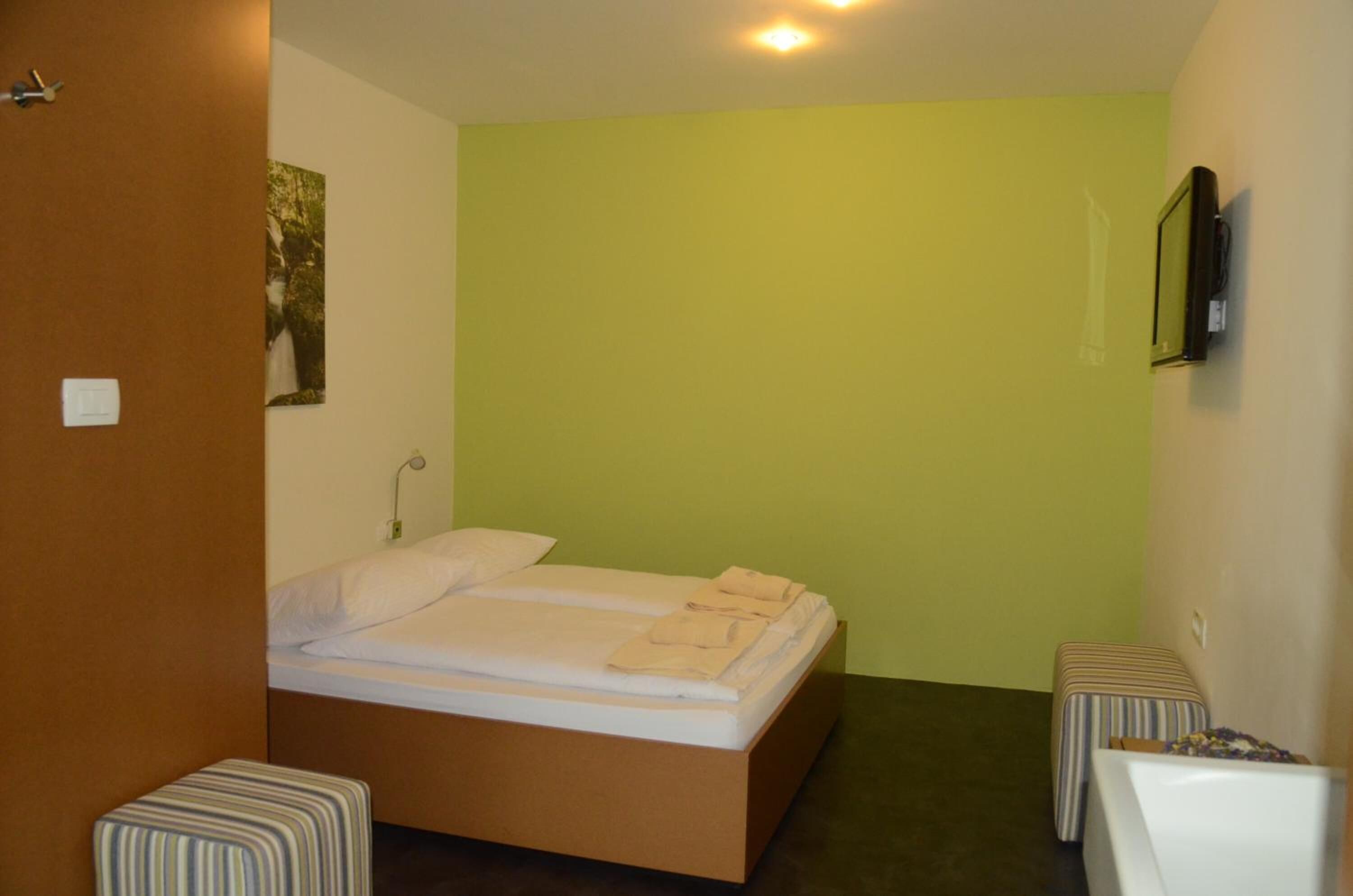 Een kleine eenpersoonskamer met een groene muur, een houten deur en een klein nachtkastje met een gestreept kussen.