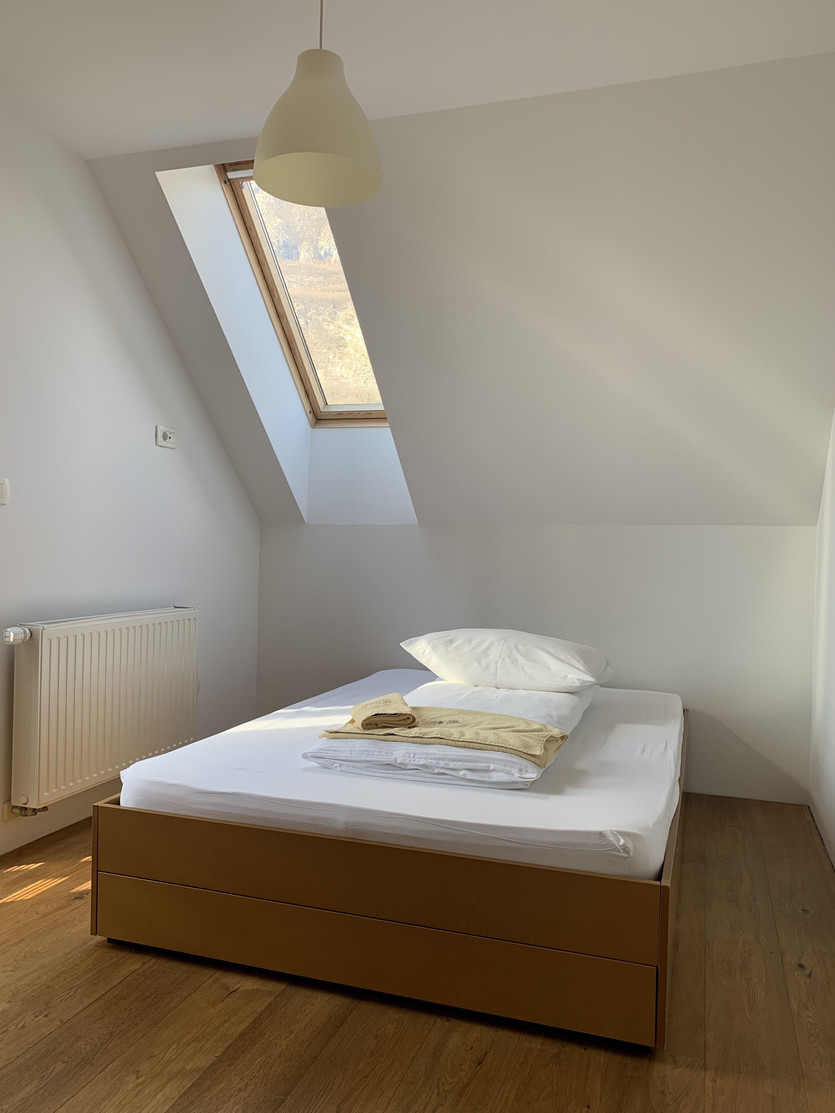 Ein Schlafzimmer mit geneigter Decke, einem Oberlicht, einem Bett mit weißer Bettwäsche, einem Holzboden und einem weißen Heizkörper.