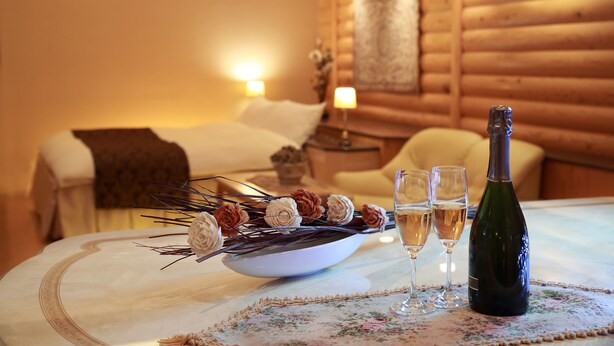 A hotel room with a bed, a bedside table with a lamp, a floral tablecloth, a bowl of dried flowers, two glasses of wine, and a bottle of wine. - Fine Garden Umeda (Osaka)