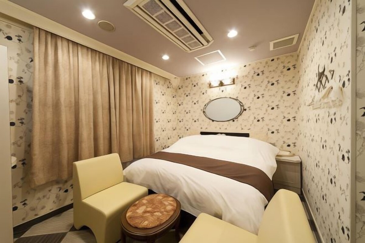 A hotel room with a bed, a bedside table, a chair, a small stool, and a mirror on the wall.