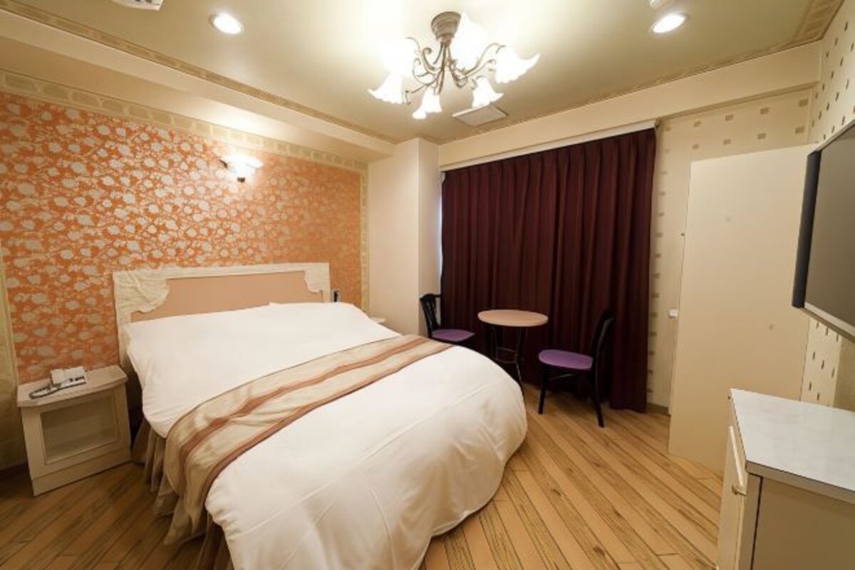 Habitación de hotel con cama, una mesita, dos sillas, televisión e iluminación en el techo.