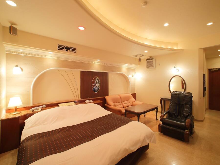 A hotel room with a large bed, a sofa, a small table, a leather chair, and a mirror.