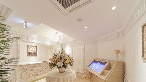 Un vestíbulo de hotel con un mostrador de recepción de mármol, una mesa redonda con un centro de mesa floral, un quiosco de información digital y una araña.