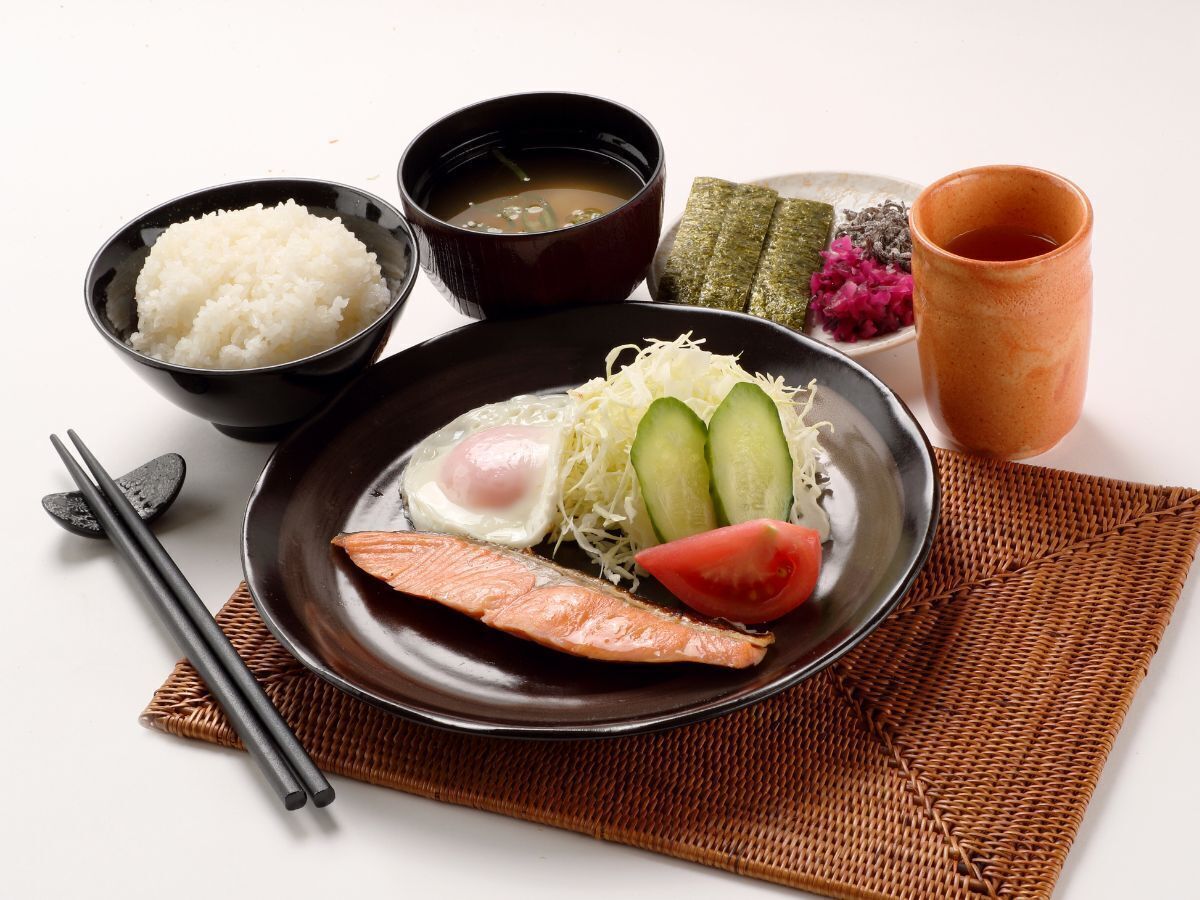 A plate with salmon, egg, cucumber, and tomato, accompanied by bowls of rice, soup, and pickled vegetables.