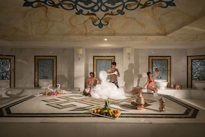 Une salle de bain luxueuse avec un sol en marbre, des appliques murales décoratives et un espace salon central agrémenté d’un arrangement de fruits.