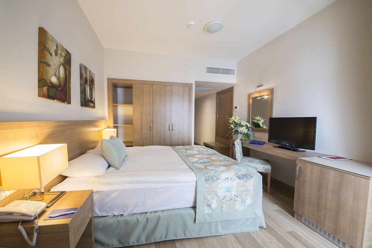 A hotel room with a bed, bedside table with a lamp, a desk with a computer, a TV, and a wardrobe.